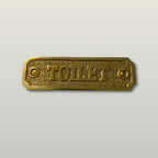 A rectangular brass toilet sign with a textured beaded border and the word 'TOILET' embossed in the center, shown on a light gray background.
