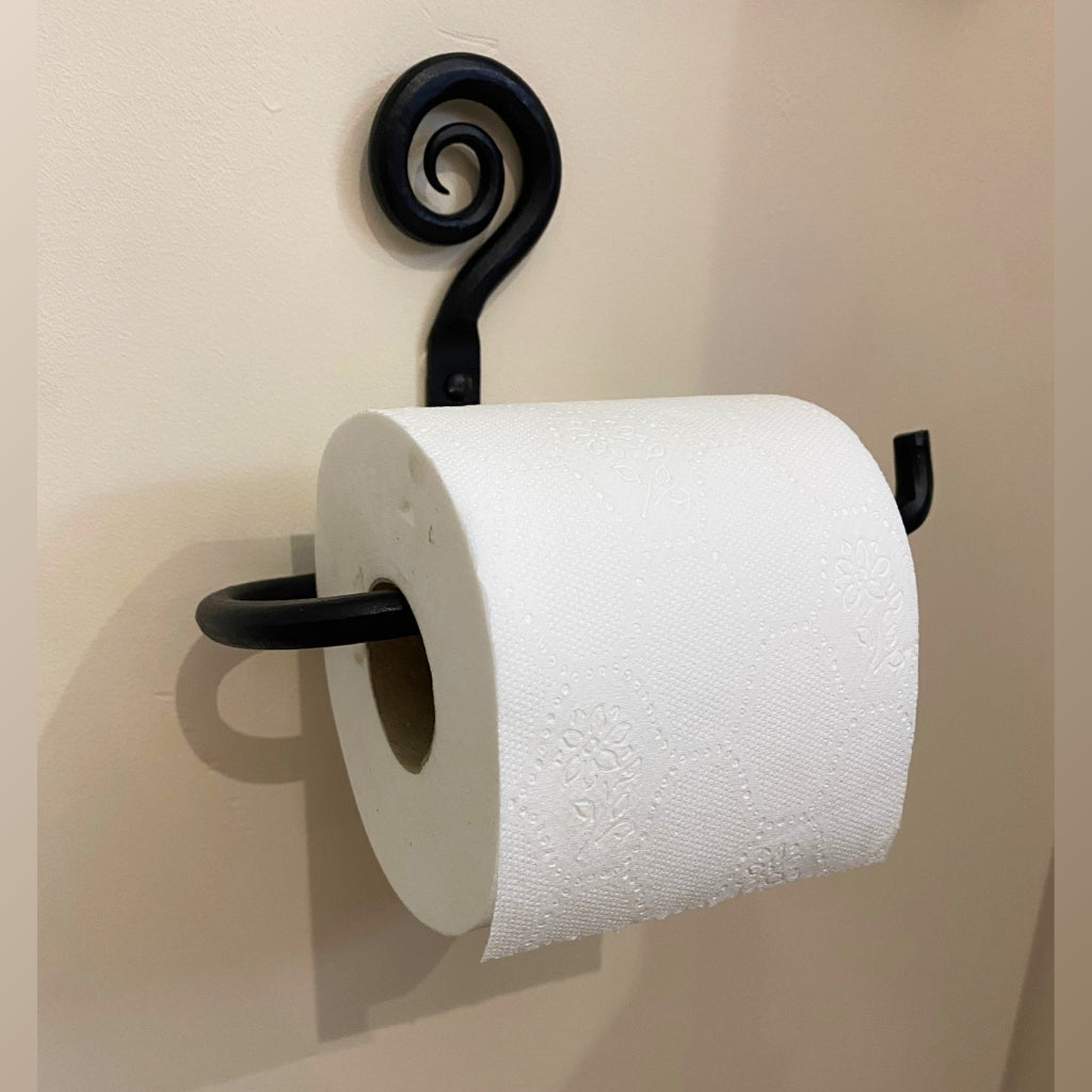 A black metal toilet paper holder for sale with a decorative scroll design on a beige wall, holding a roll of white toilet paper.