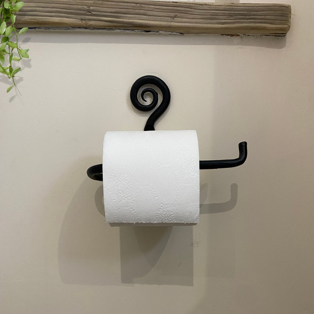 A black metal rustic toilet roll holder with a decorative spiral and a single hook arm, holding a white toilet paper roll against a beige wall.