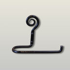 A dark, rustic hand-forged metal hook with a spiral top and a flat horizontal arm, shown against a light beige background.